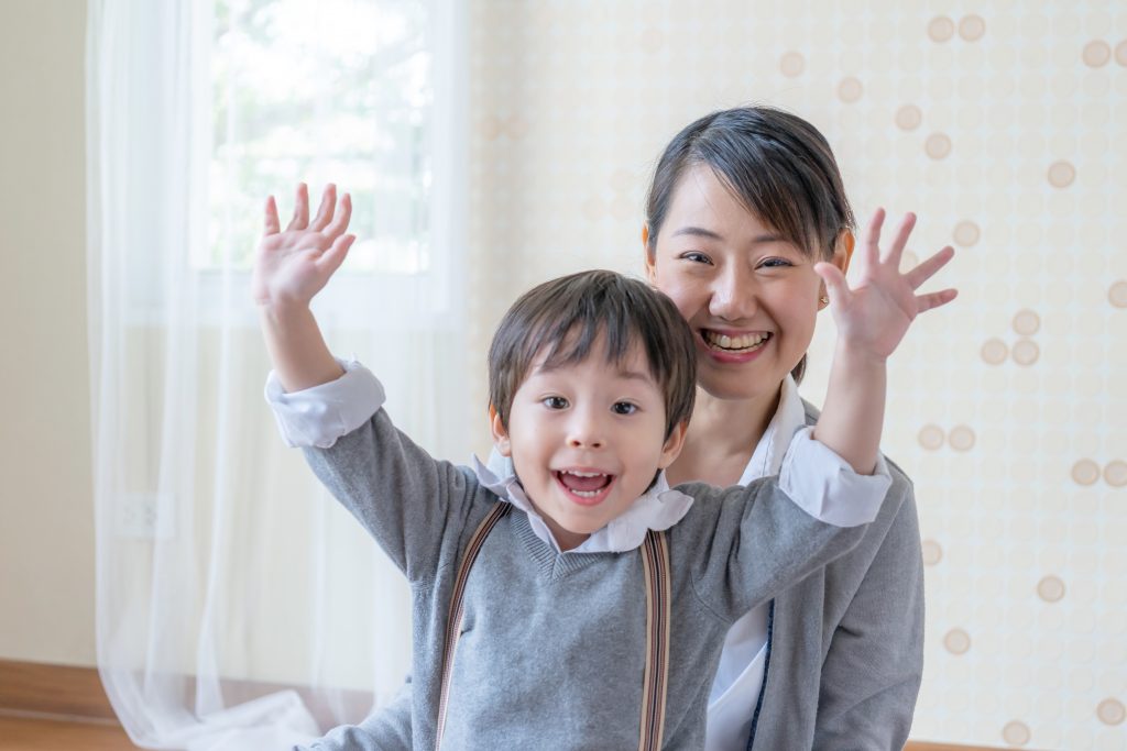 Little cute son toothy and rraise two hands above his head, young mother sitting behind him, They smiling with happily at home, copy space