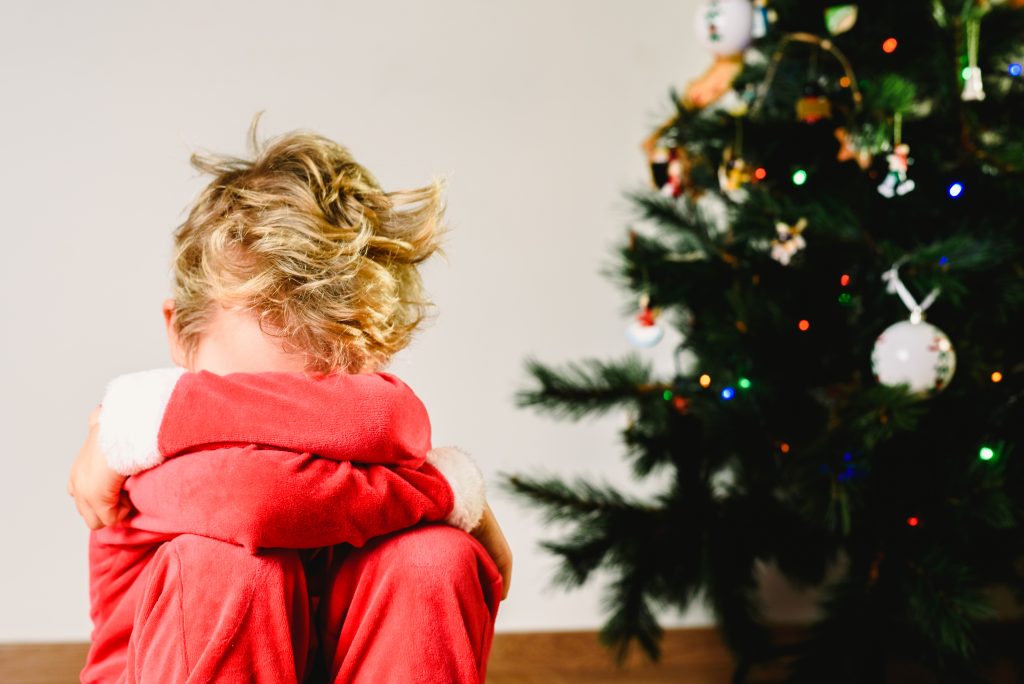 Child with disguise, anxious and sad on Christmas day with an angry face next to the Christmas tree while waiting for Santa